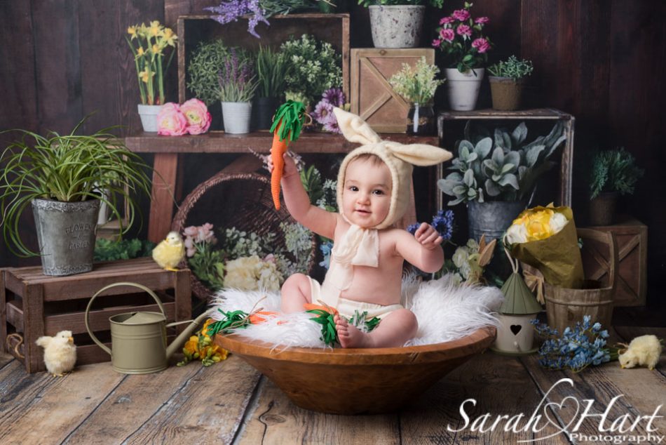 Infant in a bunny outfit holding a carrot at spring photoshoot with dark wooden backdrop